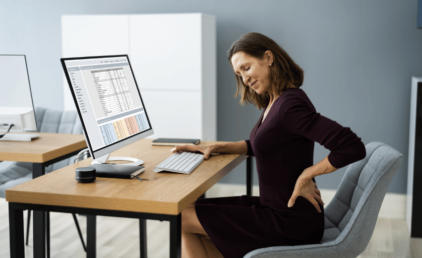 Mulher sentada em frente a uma escrivaninha em frente a um computador, expressando dor nas costas.