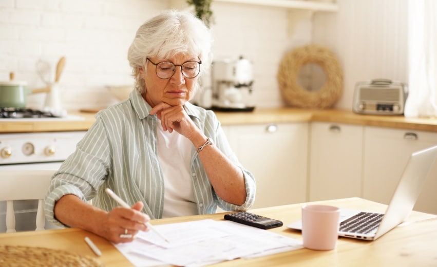 Mulher branca idosa pensativa está sentada em uma mesa enquanto observa documentos para o seu planejamento previdenciário.