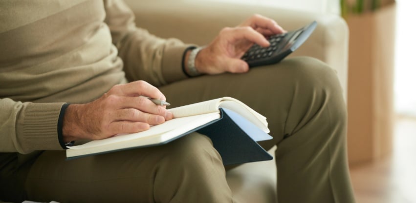A foto foca na parte inferior de um homem branco idoso sentado emm uma poltrona com um caderno em seu colo, uma calculadora em uma das mãos e uma caneta na outra, fazendo cálculos para o seu planejamento previdenciário.
