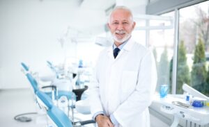 Homem branco de barba sorrindo vestindo um jaleco branco. Atrás há cadeiras de dentistas