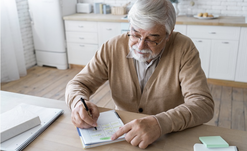Homem idoso branco escrevendo algo em um caderno realizando seu planejamento previdenciário.
