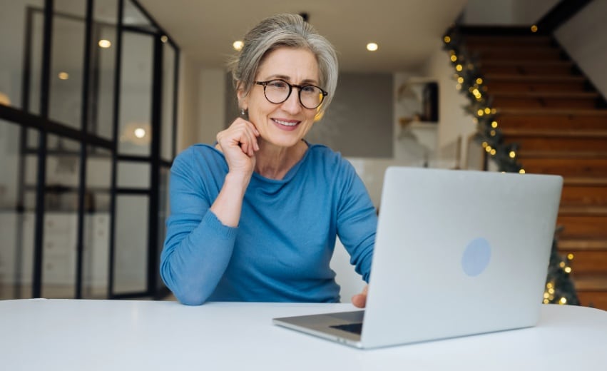 Mulher idosa branca com óculos olha sorridente para a tela de um notebook.