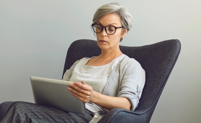 Mulher branca com cabelos curtos e grisalhos, usando óculos de grau com armação preta. Sentada em uma poltrona preta, observa um tablet.