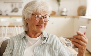 Mulher idosa com cabelos curtos e grisalhos e óculos de grau observa a tela de um celular com um semblante tranquilo.