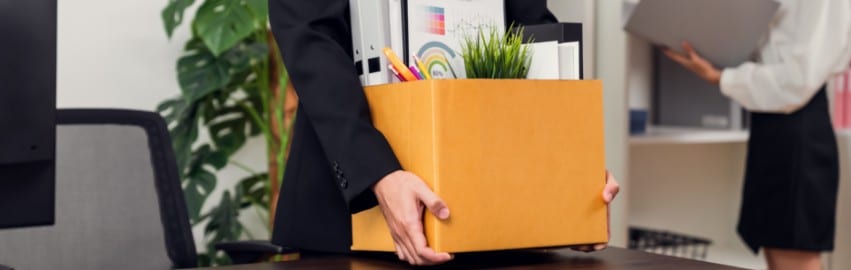 Foto de uma pessoa de terno preto carregando uma caixa de papelão com pertences dentro. Dando a entender que foi uma demissão por justa causa