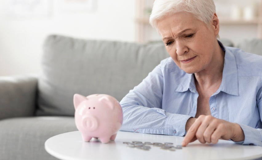 Mulher branca idosa conta moedas em cima da mesa com um semblante de preocupação. Próximo às moedas há um cofre de porquinho
