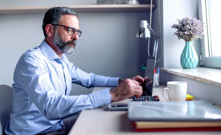 Homem branco sentado em frente a uma escrivaninha observando e manuseando um notebook.