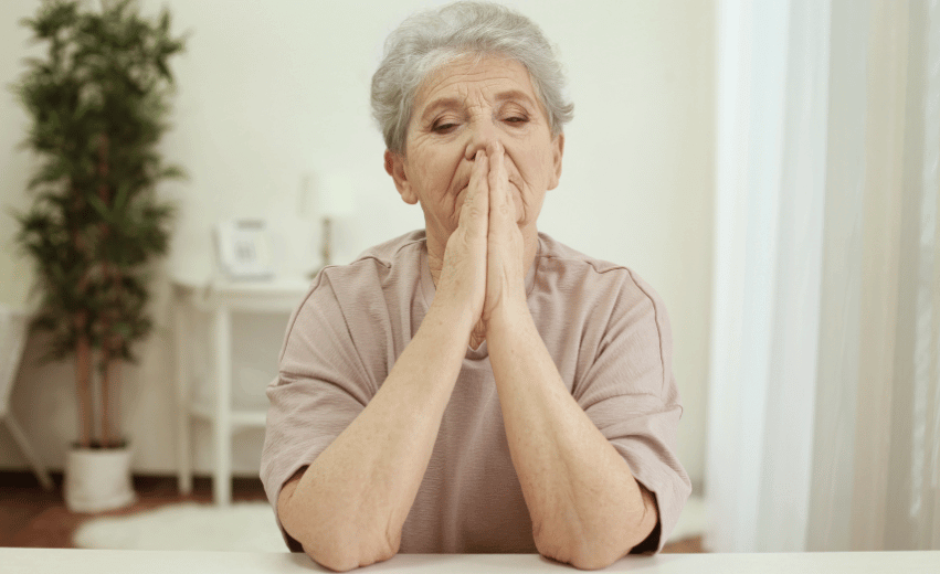 Mulher idosa com semblante preocupado e com as mãos a frente em posição de oração.