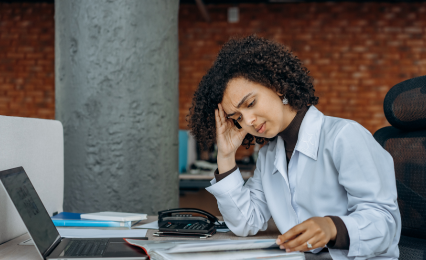 Mulher negra com cabelos cacheados sentada em frente a uma escrivaninha. Ela aparenta estar esgotada, um dos sintomas da síndrome de burnout.